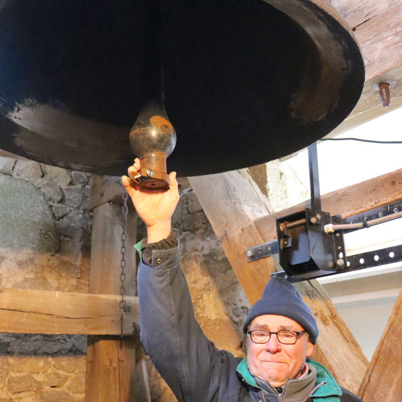 Bernard tenant le battant de Saint-Pierre, la plus grosse cloche de l&rsquo;&eacute;glise Saint-Pierre-&egrave;s-Liens de Campuac. Derri&egrave;re lui, le bo&icirc;tier noir est le syst&egrave;me &eacute;lectrique qui actionne le marteau pour frapper la cloche et sonner les heures. - Aline Amodru-Dervillez
